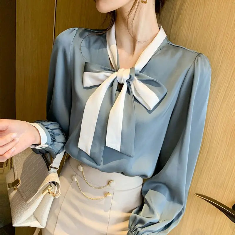 Woman wearing a light Blue blouse with a large White bow, standing against a wooden wall.