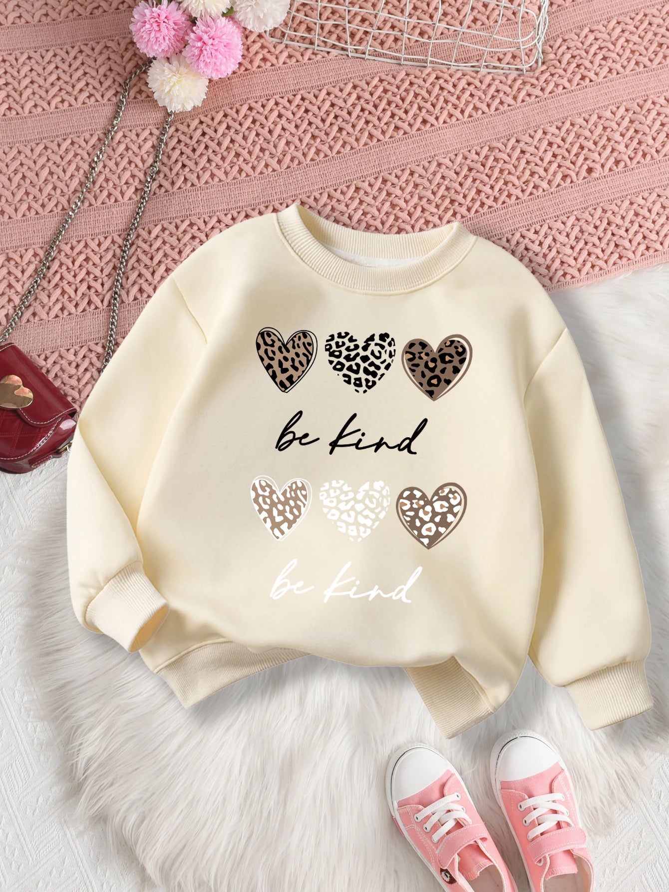 Beige sweatshirt with heart designs and 'be kind' text on a Pink textuRed surface.