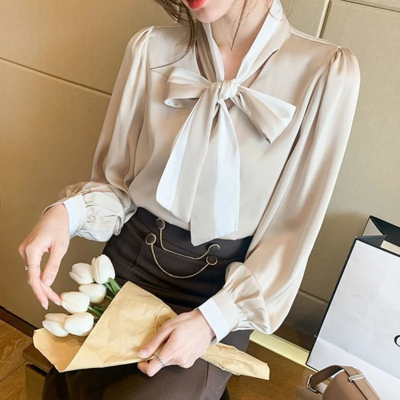 Woman wearing a beige blouse with a bow tie, holding flowers and a gift bag.