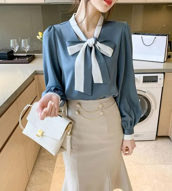 Woman in a kitchen wearing a Blue blouse with a White bow and beige skirt.