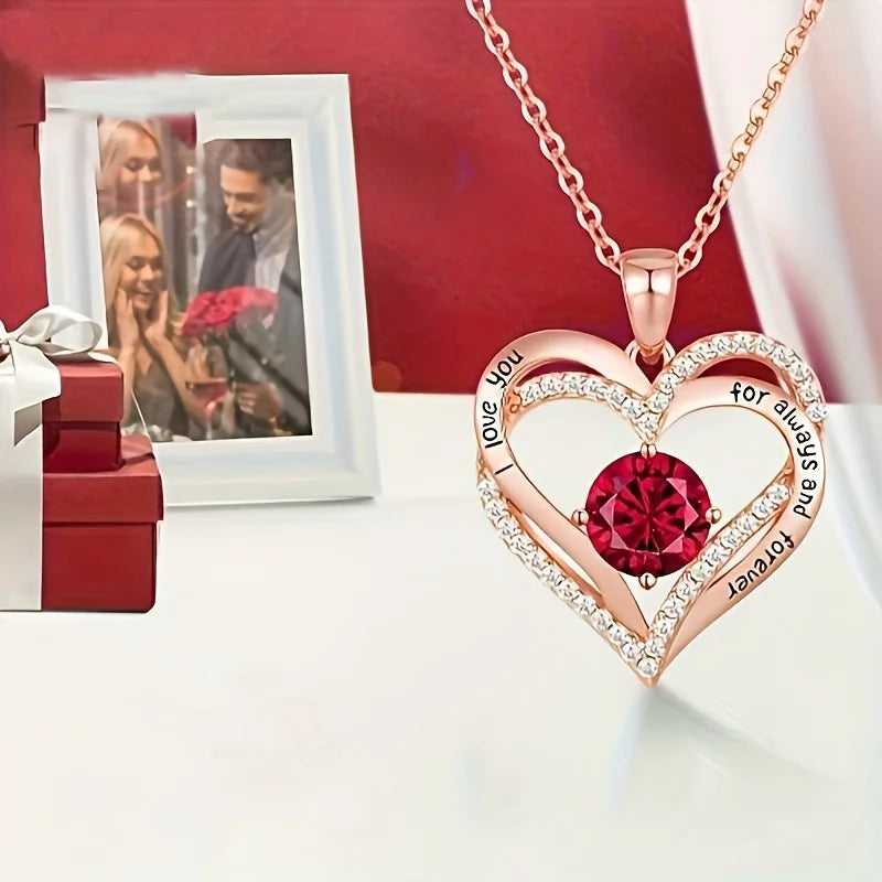 Heart-shaped gold necklace with a Red gemstone and engraved text, placed on a White surface with a Red background.