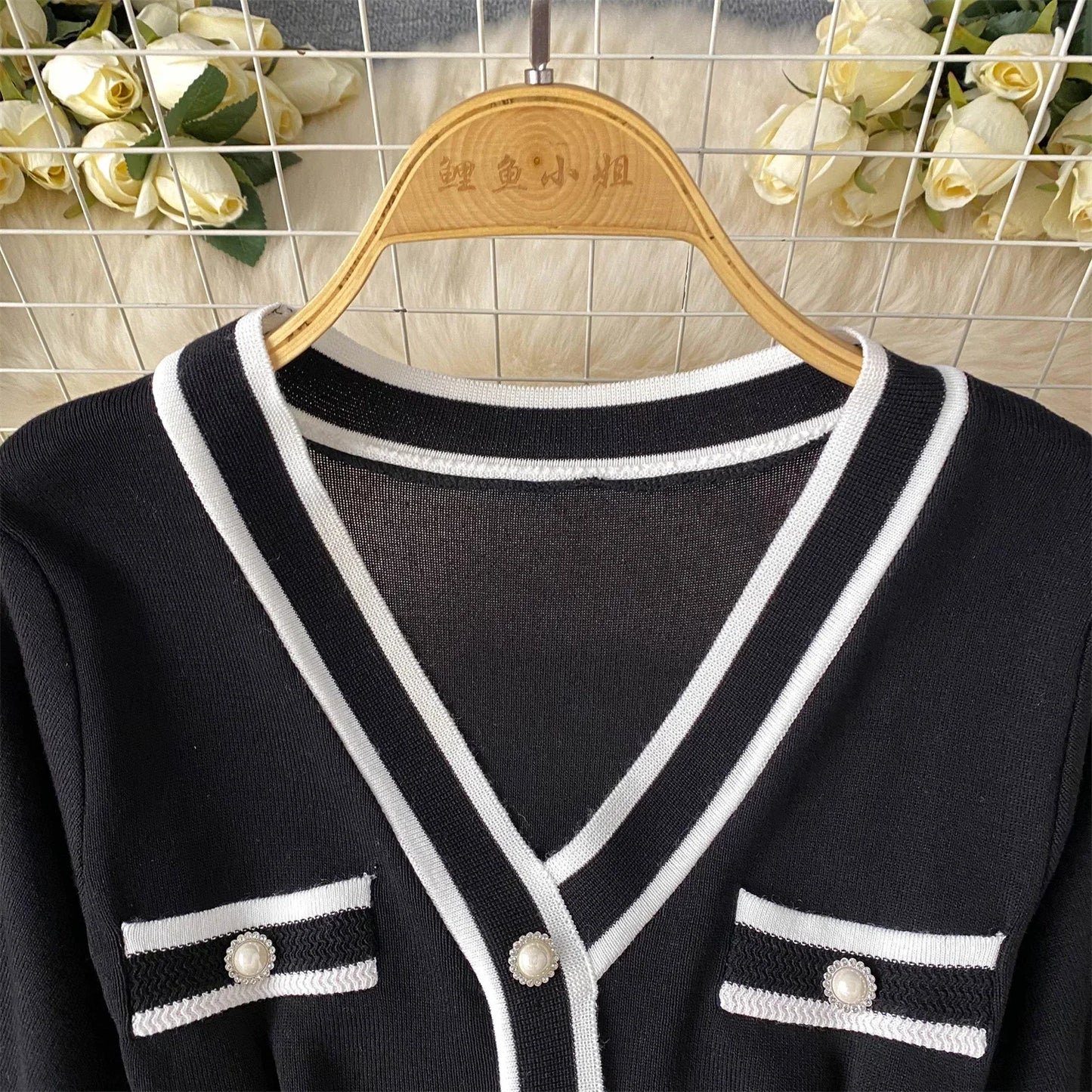Black cardigan with White trim on a wooden hanger against a tiled wall background.