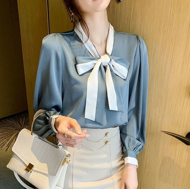 Woman wearing a light Blue blouse with a White bow tie, holding a beige handbag.